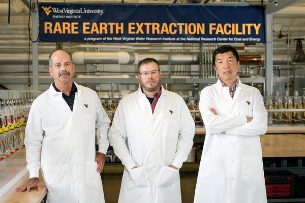 Three men in lab coats stand in front of the Rare Earth Extraction Facility sign