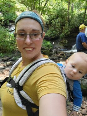 A person wears a blue headband and glasses along with backpack that is holding a baby. The adult is in a yellow shirt. The baby is in a blue shirt.