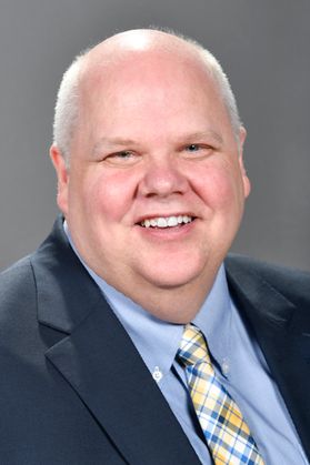 A headshot of WVU employee Jack Thompson. He is pictured against a gray background wearing a navy blue coat over a light blue shirt and a blue and yellow plaid tie. 