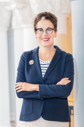 This is a portrait of Provost Maryanne Reed. She has her arms crossed and is wearing a blue suit jacket, blue and white striped top, dark rimmed glasses and is standing in front of several white columns.