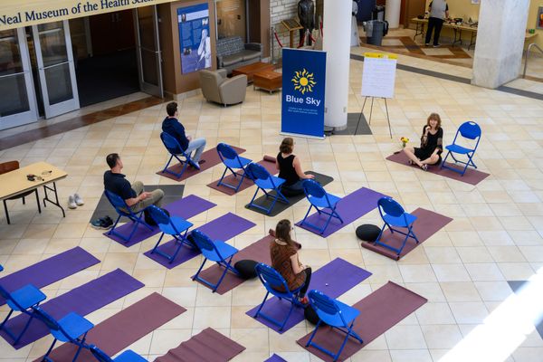A photograph showing a yoga class. A dozen maroon yoga mats are situated in a grid pattern on the floor. Each mat also has a blue folding chair sitting on top of it. The female leader sits cross legged at the front of the group of four participants. 