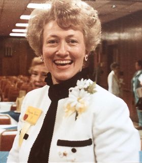 smiling woman, neatly coiffed hair, white jacket, black turtleneck top, corsage