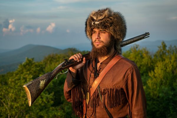 young man in buckskins, coonskin cap, musket over his shoulder, mountains in background