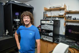 Biology professor Kevin Daly, wearing a blue shirt, is pictured standing in his lab at WVU with two large pieces of equipment behind him. .