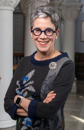 Headshot of  WVU provost Maryanne Reed. She is standing inside with white columns in the background. She is wearing a gray, floral patterned sweater and circle framed glasses. She has short, salt and pepper hair. 