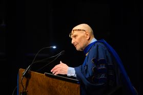 Dr. Michael Apuzzo leans on a wooden podium while wearing a blue robe. He has glasses on top of his head.