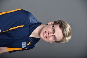 A person with short blond hair and glasses in a gold and blue shirt with Statler College on the left side smiles at the camera