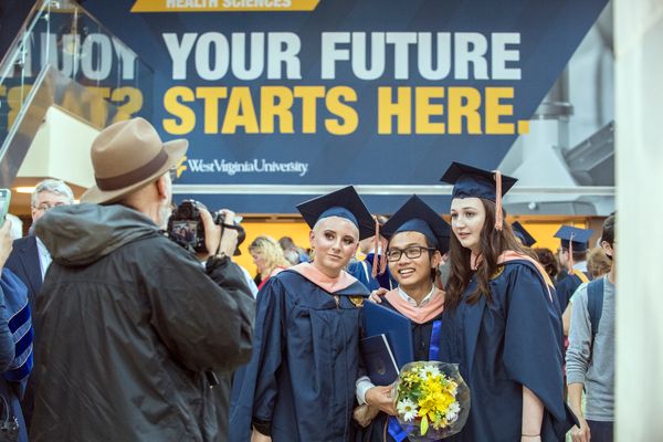 Man in a brown hat takes a picture of smiling students wearing blue caps and gowns on graduation day