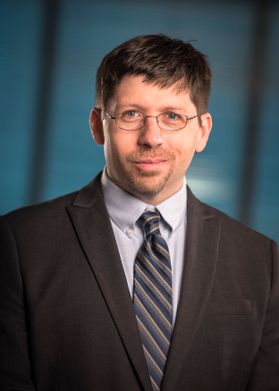 Headshot of Brian Lego. He is standing inside in front of blue glass background. He is wearing a charcoal sport coat with a striped tie and light colored shirt. His hair is brown and short. He has a goat tee and wears glasses. 