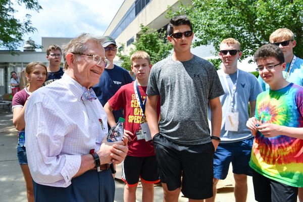 President Gordon Gee, wearing a plaid shirt and purple tie, smiles while talking to a group of students while standing outside chatting