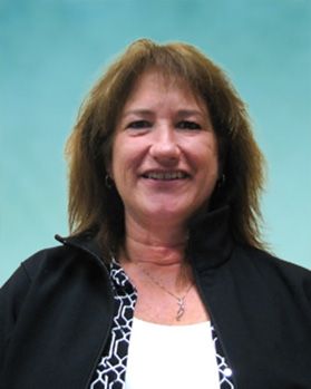 Headshot of WVU program manager Mary Gibbs. She is pictured in front of a light blue wall wearing a white shirt with a black and white patterned jacket on top. She has shoulder length brown hair. 
