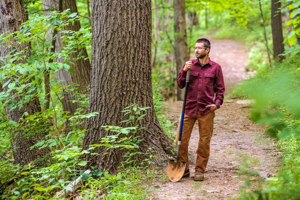 Man stands in forest in red flannel and brown pants holding a shovel on a trail with green leaves around him 