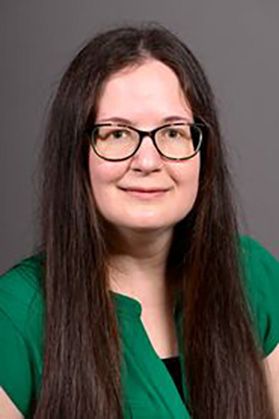 Headshot of WVU researcher Katie Corcoran. She is pictured against a gray wall and is wearing a kelly green blouse. She has long, straight brown hair and is wearing glasses. 