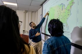 WVU professor Omar Abdul-Aziz points to a large map on the wall while talking to two of his graduate student. You can only see the back of their heads in the photo. Abdul-Aziz is wearing khakis and a blue sweater. 