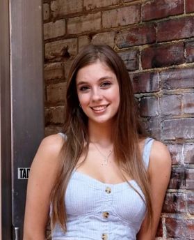 Headshot of WVU student Madyson Kimble. She is pictured leaning against a brick wall. She is wearing a white, button up blouse and her long brown hair is cascading down the front of her. 