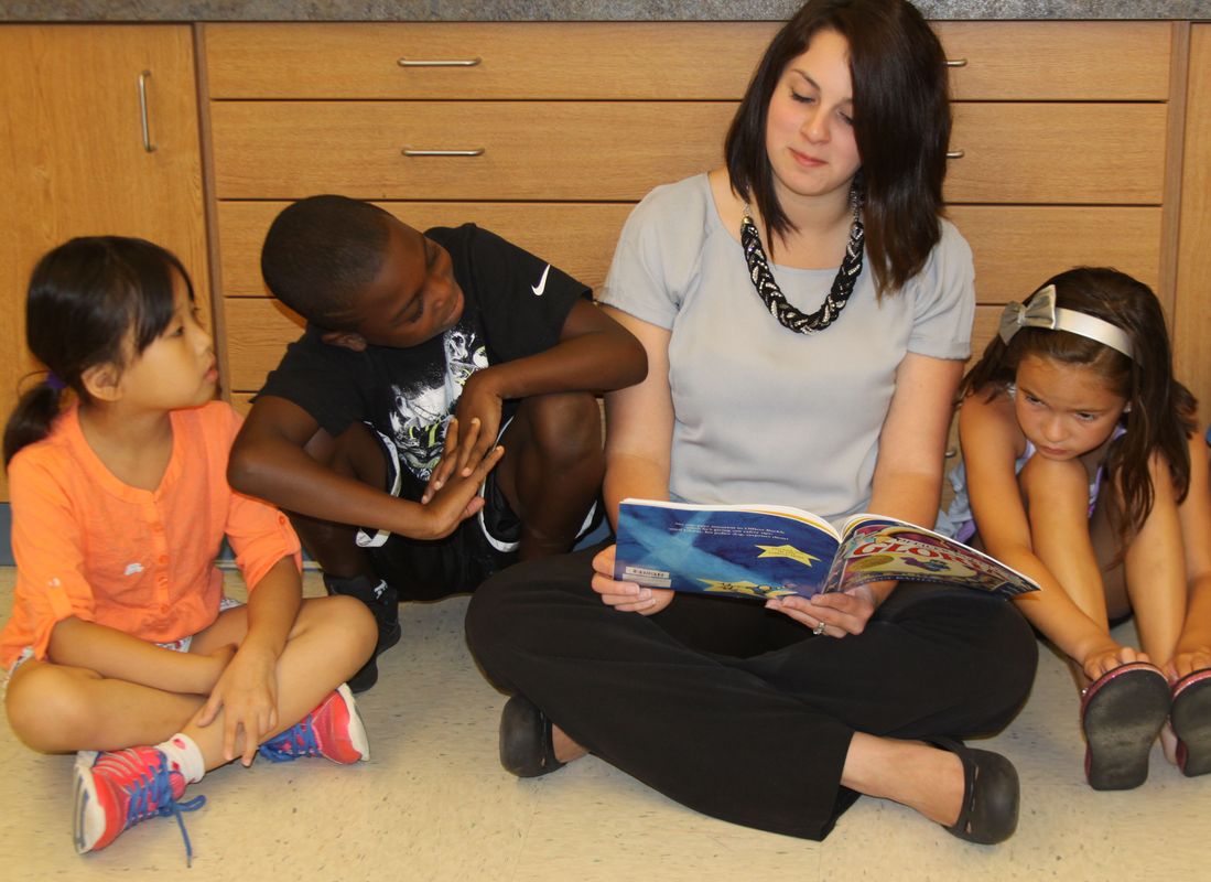 A teacher in a gray top with a thick necklace reads a book to three young students while leaning against a brown wall.