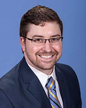 Headshot of WVU researcher Jonathan Herczyk. He is pictured against a blue background and is wearing a dark colored suit jacket with a white dress shirt and a blue and gold striped tie. He has short brown hair and wears glasses. 