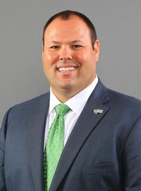 This is portrait of Wren Baker, the new director of Athletics at West Virginia University. He is wearing a dark suit and green tie in front of a gray background.
