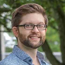 Headshot of WVU researcher Nicholas Szczecinski. He is standing outside with trees in the background and is wearing a light blue patterned shirt. He has short light colored hair and beard and wears black framed glasses. 