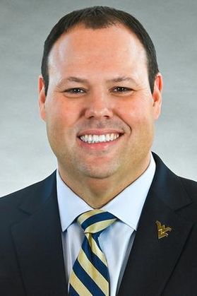 A man smiling wearing a suite with a WVU pin and a gold and blue stripped tie.