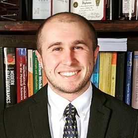 Photograph of WVU professor Thomas Steinberger. He is pictured standing in front of a bookshelf wearing a dark coat, white dress shirt, and dark patterned tie. He has shaved hair and a bright smile. 