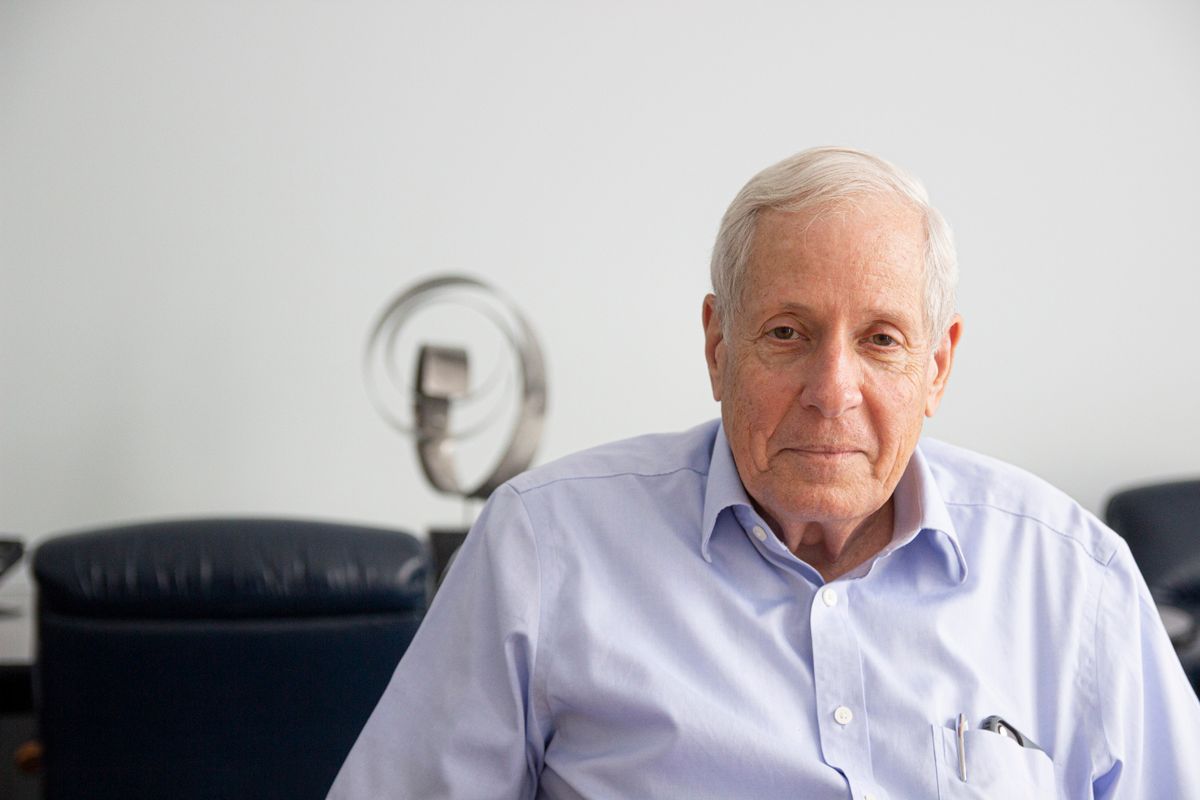 Older gentleman in a blue button down poses for a headshot with a couch and a sculpture in the background.