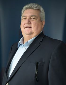 A person wearing a dark suit jacket and light blue button up shirt with short gray hair stands in front of a dark background.