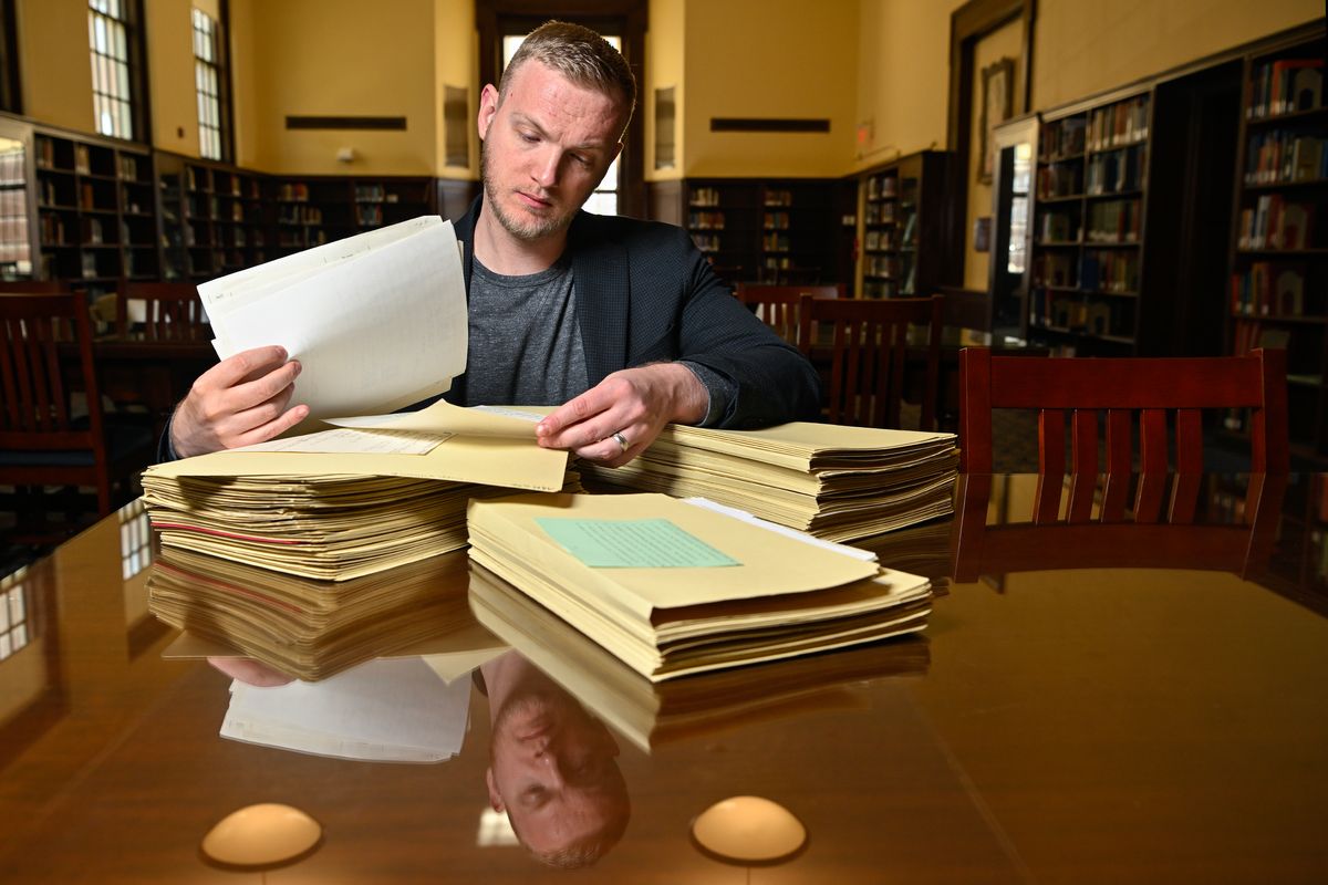 WVU researcher Jonathan King sifts through stacks of papers on a large wooden table for his research. He is seated in a library and stacks of books can be seen in the background. 