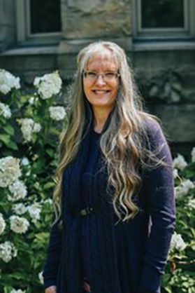 Photograph of WVU professor Melanie Page. She is pictured outside with a building behind her. There are also bushes full of white flowers on either side of her. She has long light colored hair and is wearing classes and a dark colored shirt. 