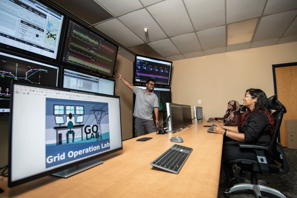 Three computer science and electrical engineering students discuss a power grid simulation project. Two are seated at a table looking at a wall full of monitors. The third is standing pointing at one of the screens. 