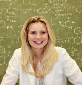 Headshot of WVU student Courtney Glenn. She is pictured standing in front of a chalk board full of chemistry equations. She is wearing a white lab coat and has long blonde hair. 