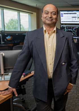 Headshot of WVU Professor Anurag Srivastava. He is standing in his lab wearing a dark gray suit and yellow dress shirt. He has one hand on his hip and the other on a table next to him. 