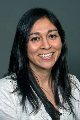 Headshot of WVU faculty member Megan Govindan. She is pictured in front of a blue gray background wearing a white blouse. She has long, black hair. 
