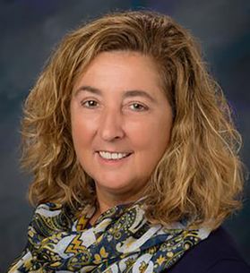 Headshot of WVU alumni Tracy Schoenadel. She is pictured against a dark blue gray background. She is wearing a dark colored top with a WVU branded scarf wrapped around her neck. She has shoulder length blond hair. 