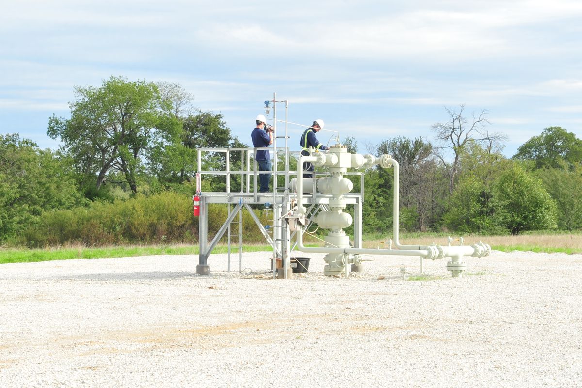 two men in hardhats and coveralls stand on gas well structure