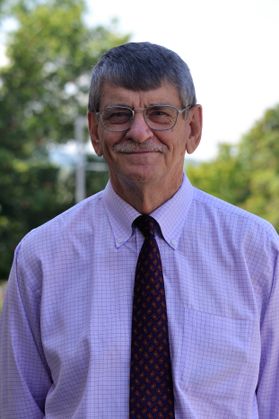 Headshot of WVU researcher Ian Rockett. He is pictured outside wearing a checked shirt with a dark colored tie. He is an older gentleman with gray hair and glasses. 