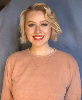 Headshot of researcher Sara Starcovic. She is pictured against a blue background and is wearing a peach blouse. She has short, wavy blonde hair and red lip stick.