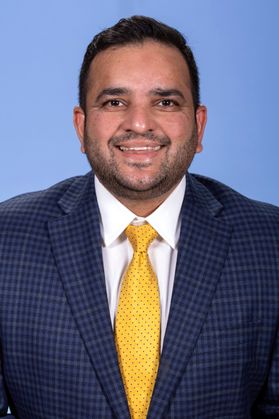 Headshot of WVU professor Shailendra Singh. He is pictured against a blue background and is wearing a blue checkered sport coat over a white dress shirt with a gold tie. He has short, black hair and a 5 o'clock shadow.  