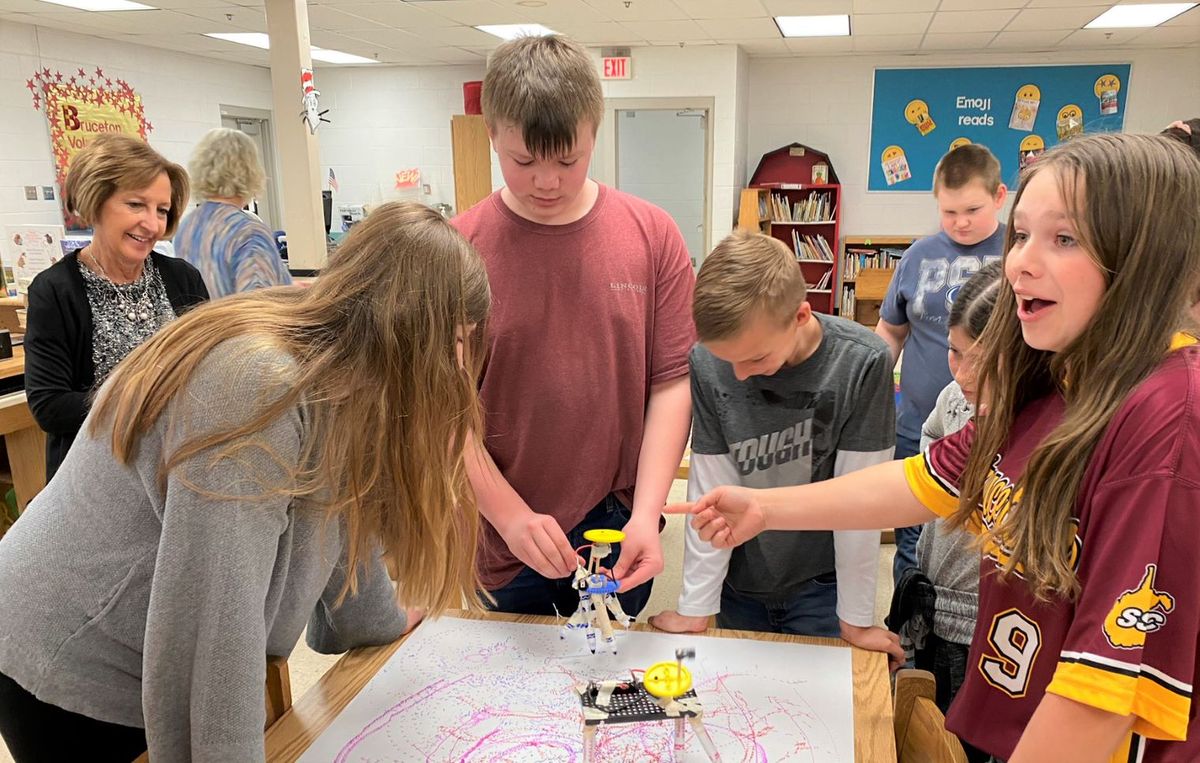 Six middle school students gather around a table in a classroom as their teacher observes. They are participating in a STEAM TAC experience that includes using small robots to color a sheet of paper with markers. 