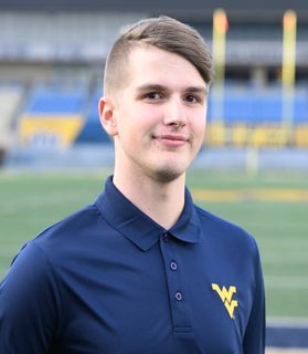 This is portrait of Braden Adkins who has short, light brown hair and is wearing a navy blue polo shirt with a Flying WV on the left side. Braden is standing on Mountaineer Field.