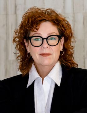 woman with red hair, large glasses, white shirt, dark jacket