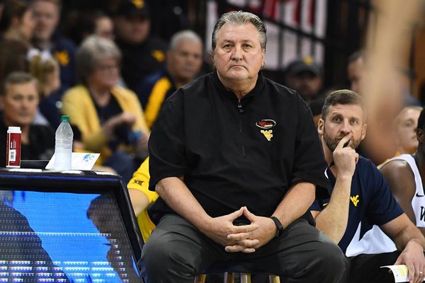 Bob Huggins sits on a stool on the basketball court wearing a black pullover shirt. His hands are clasped at his waist.