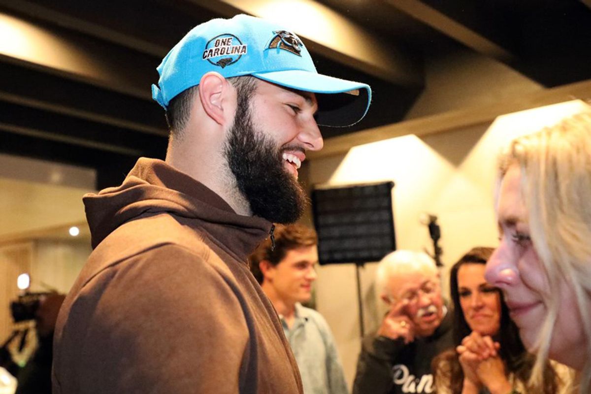 man with a dark beard wearing a light blue hat and brown hoodie smiles at a blonde woman