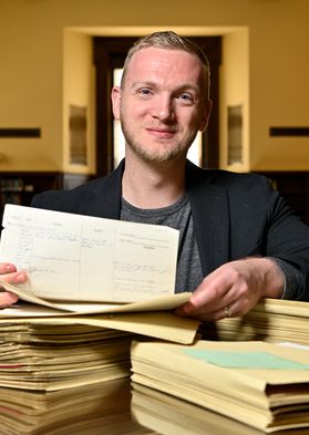 WVU researcher Jonathan King is seated at a large table in a library sifting through stacks of papers. He is wearing a dark jacket over a gray shirt and has short, light colored hair. 