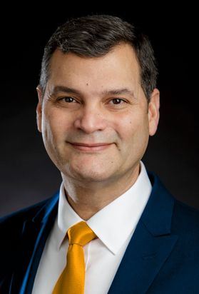 Headshot of WVU Extension Deal Jorge Atiles. Atiles is wearing a dark suit, white shirt, and bright, gold tie. He had short brown hair. 