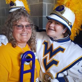 Amy Hall and her daughter Keeley Hagans at a WVU event. The mother is wearing a gold tshirt and her daughter is wearing a white WVU Marching Band uniform. Both are holding their trombones. 