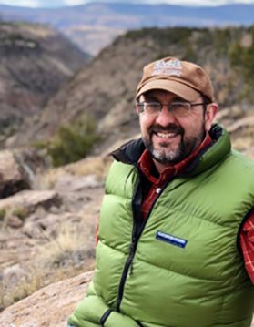 Photograph of WVU researcher William Peterjohn. He is standing outside in a rural location. He is wearing a green puffer vest over a red shirt, a brown hat and glasses. 