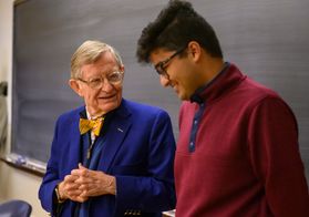 President Gordon Gee interacting with WVU student. Gordon Gee is wearing a blue suite with a bright gold tie. He has grey hair with round glasses. The WVU student is wearing a dark red sweater with round glasses, and he has dark hair.