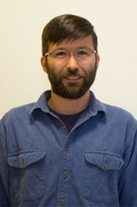 Headshot of WVU professor Zach Fowler. He is pictured against an off-white background wearing a blue button-up shirt. He has brown hair and a brown beard. He also wears glasses. 