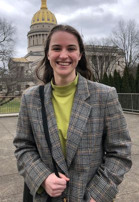 Photograph of scholarship recipient Teagan Kuzniar. She is standing in front of the West Virginia capitol building wearing a black and brown tweed patterned jacket over a lime green shirt. She has long, dark hair and has a black bag slung over her arm. 